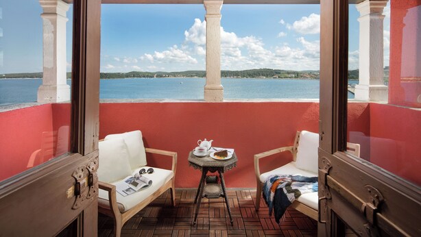 Een balkon met twee witte stoelen met kussens, een klein rond tafeltje met een theepot en een bord eten, en uitzicht op een wateroppervlak en verafgelegen land.