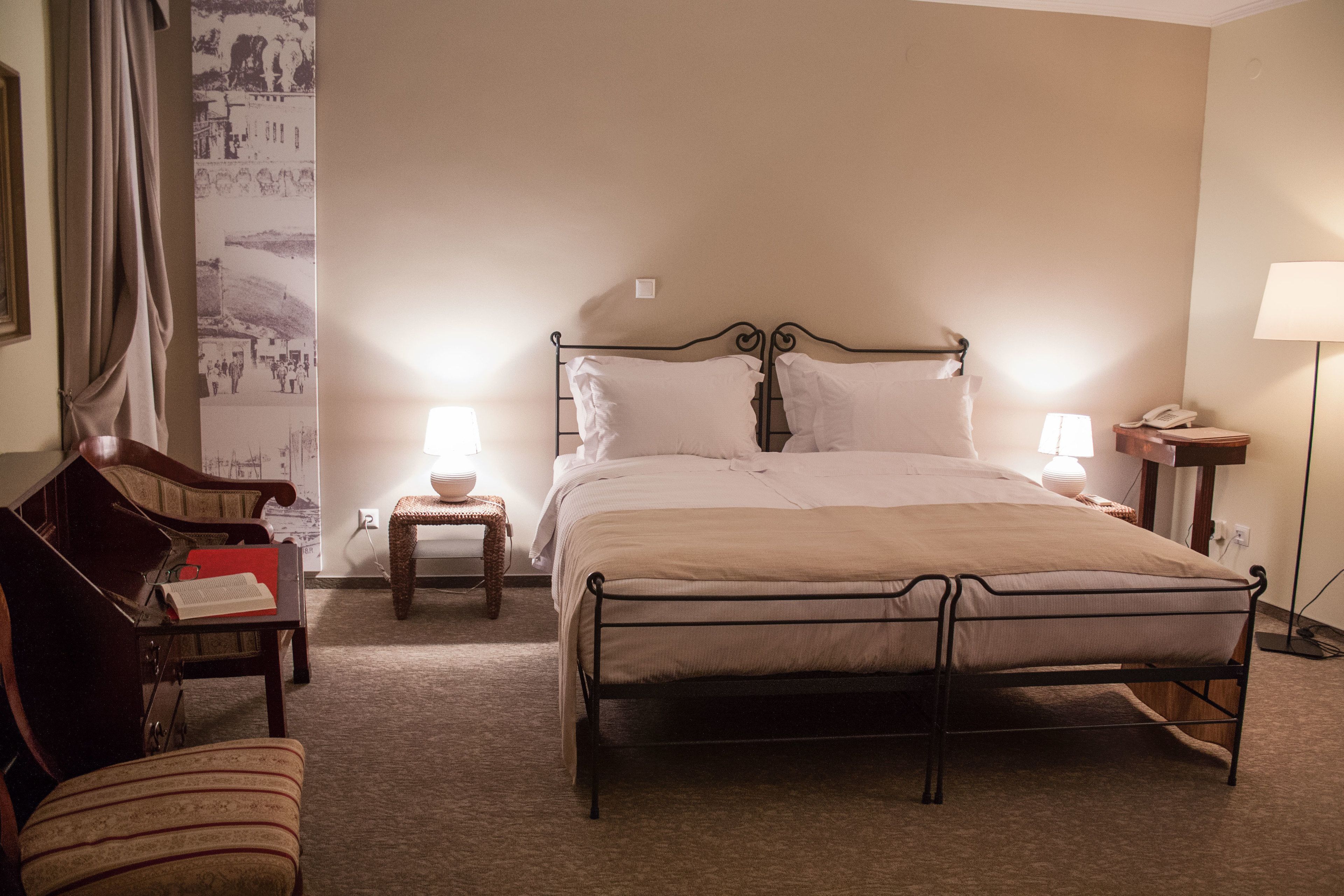 A neatly arranged hotel room with a metal bed frame, two bedside tables with lamps, a chair, and a wall with a cityscape wallpaper.