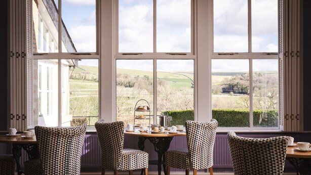 A dining area with patterned chairs and a table set for tea, overlooking a rural landscape through large windows. - The Fell Hotel (Skipton)