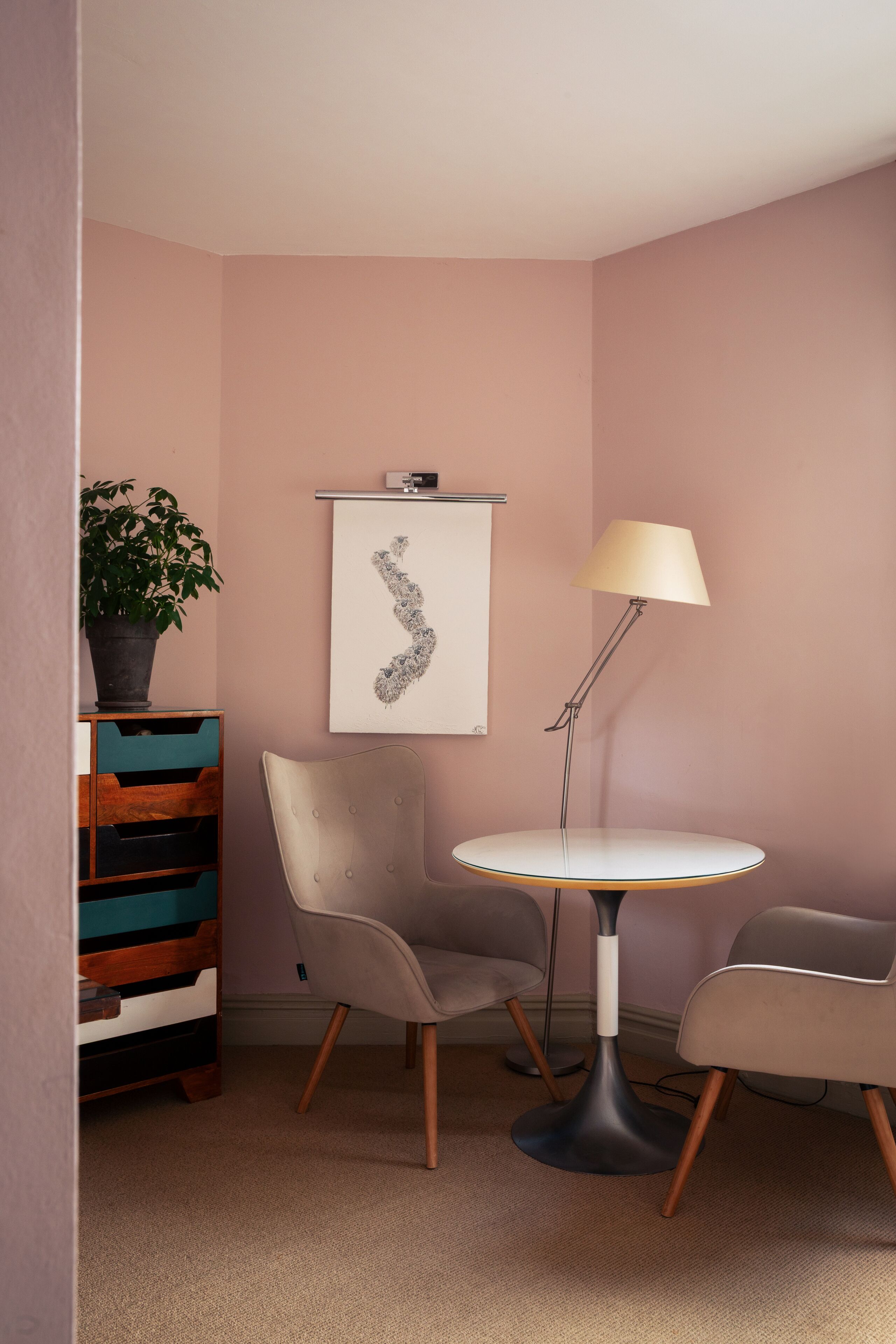 Ein Zimmer mit rosa Wand, einem runden Tisch mit Lampe, zwei Stühlen, einem Holzkasten mit Schubladen und einer Blumentopf-Pflanze.