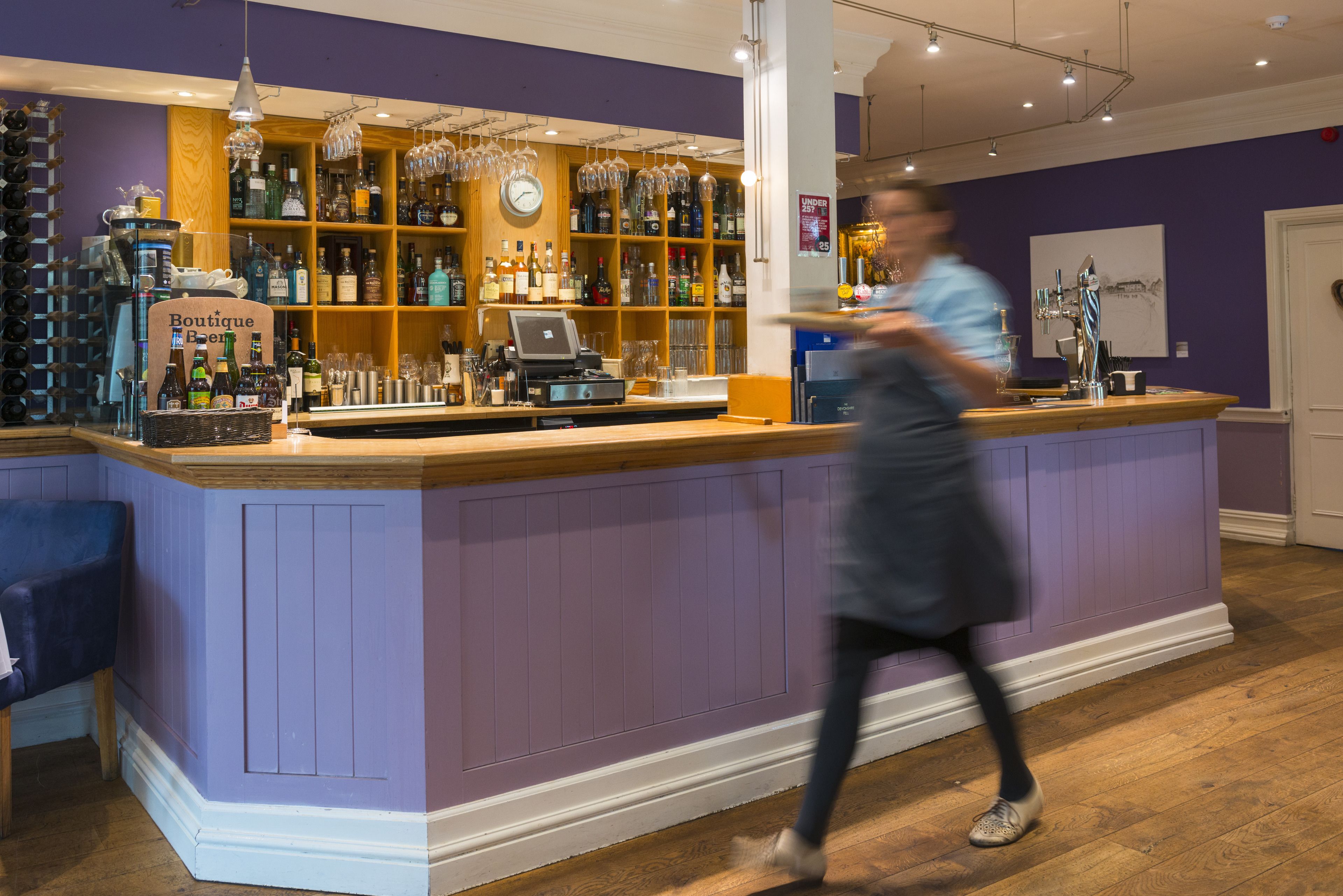A bar area with a purple wall, wooden shelves with bottles, and a cash register.