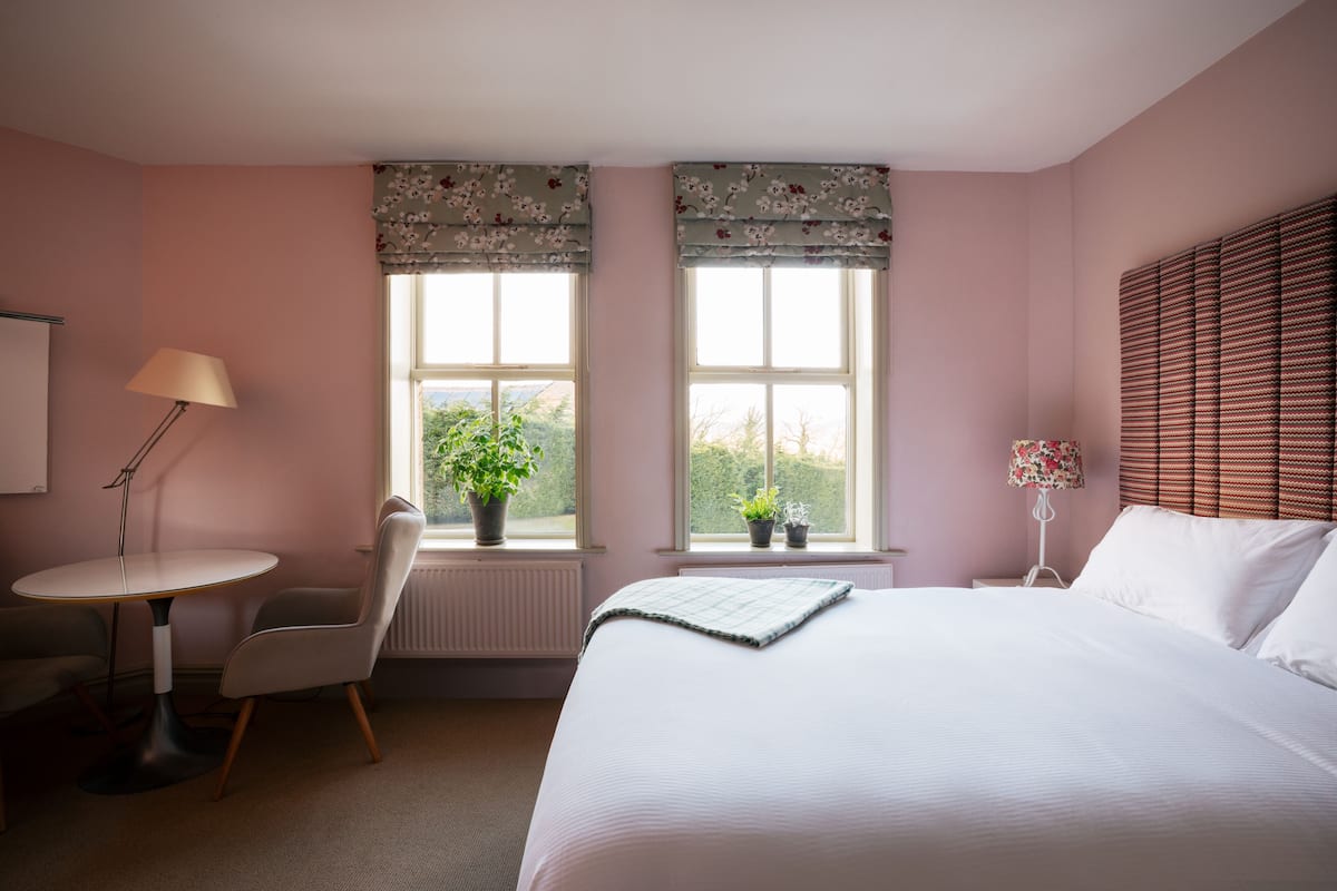 A bedroom with a bed, a chair, a small table, two windows with curtains, and a radiator.