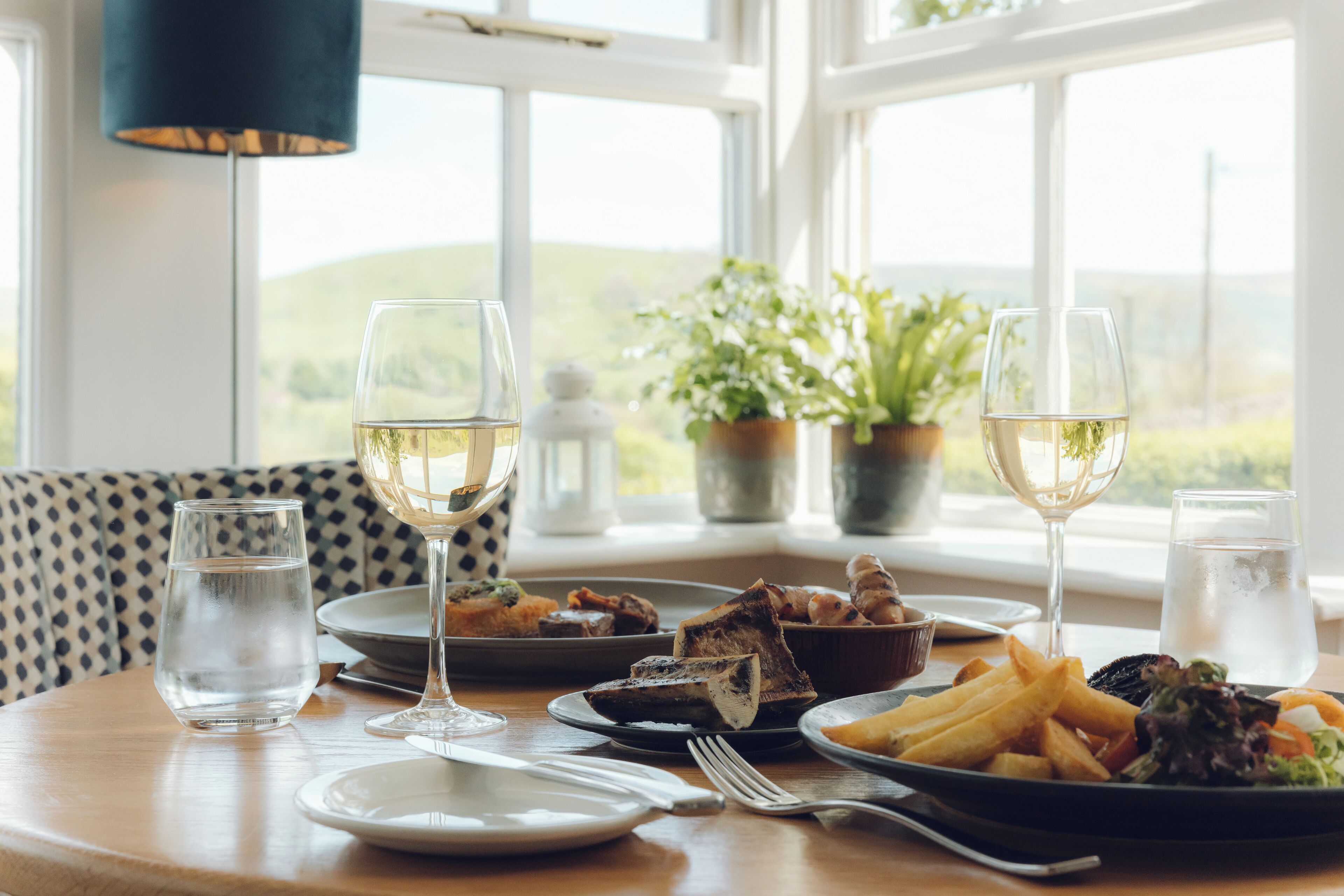 A dining table with plates of food, glasses of water and wine, and a view of the outdoors.