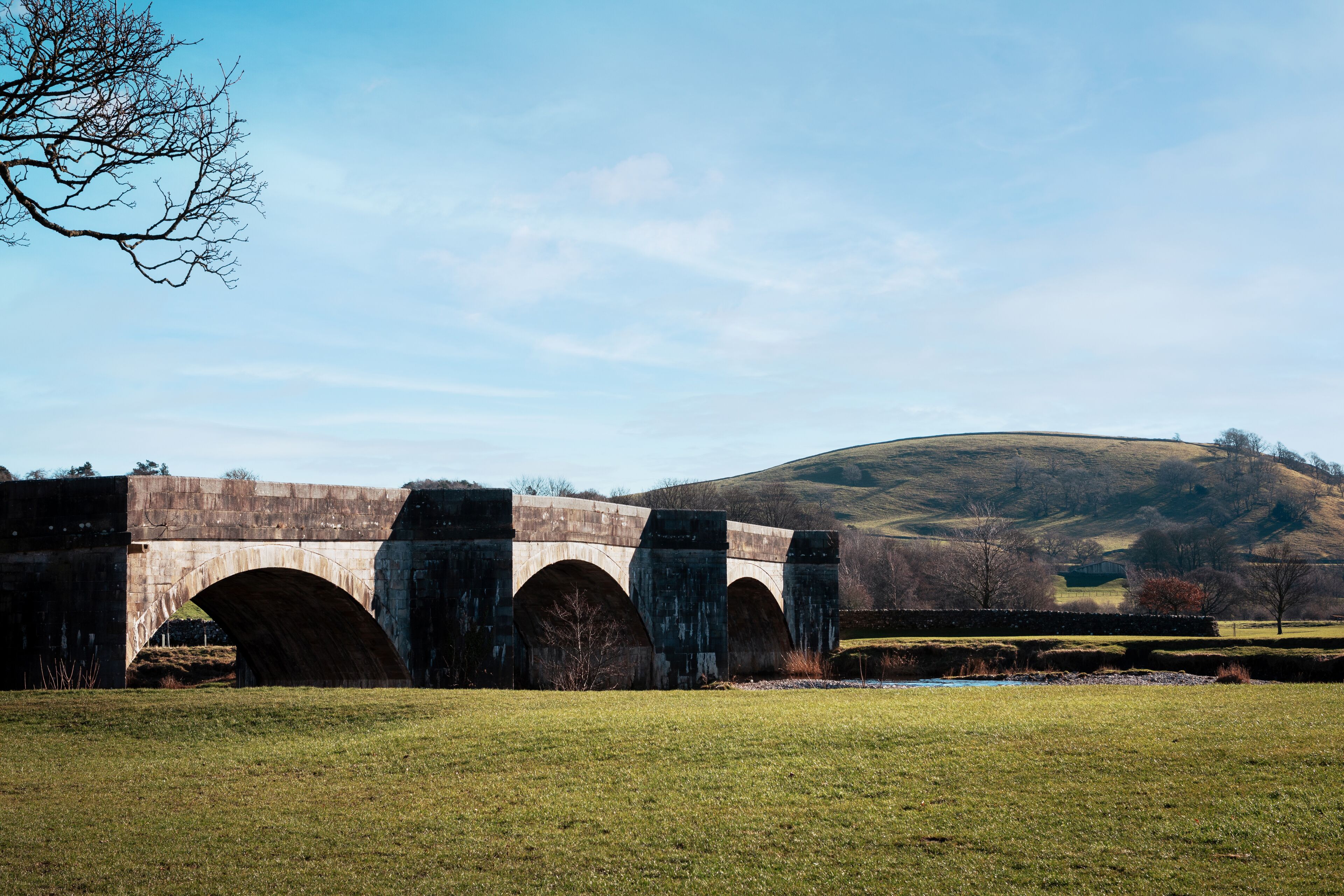 A stone bridge over a river with a grassy field in the foreground and hills in the background.