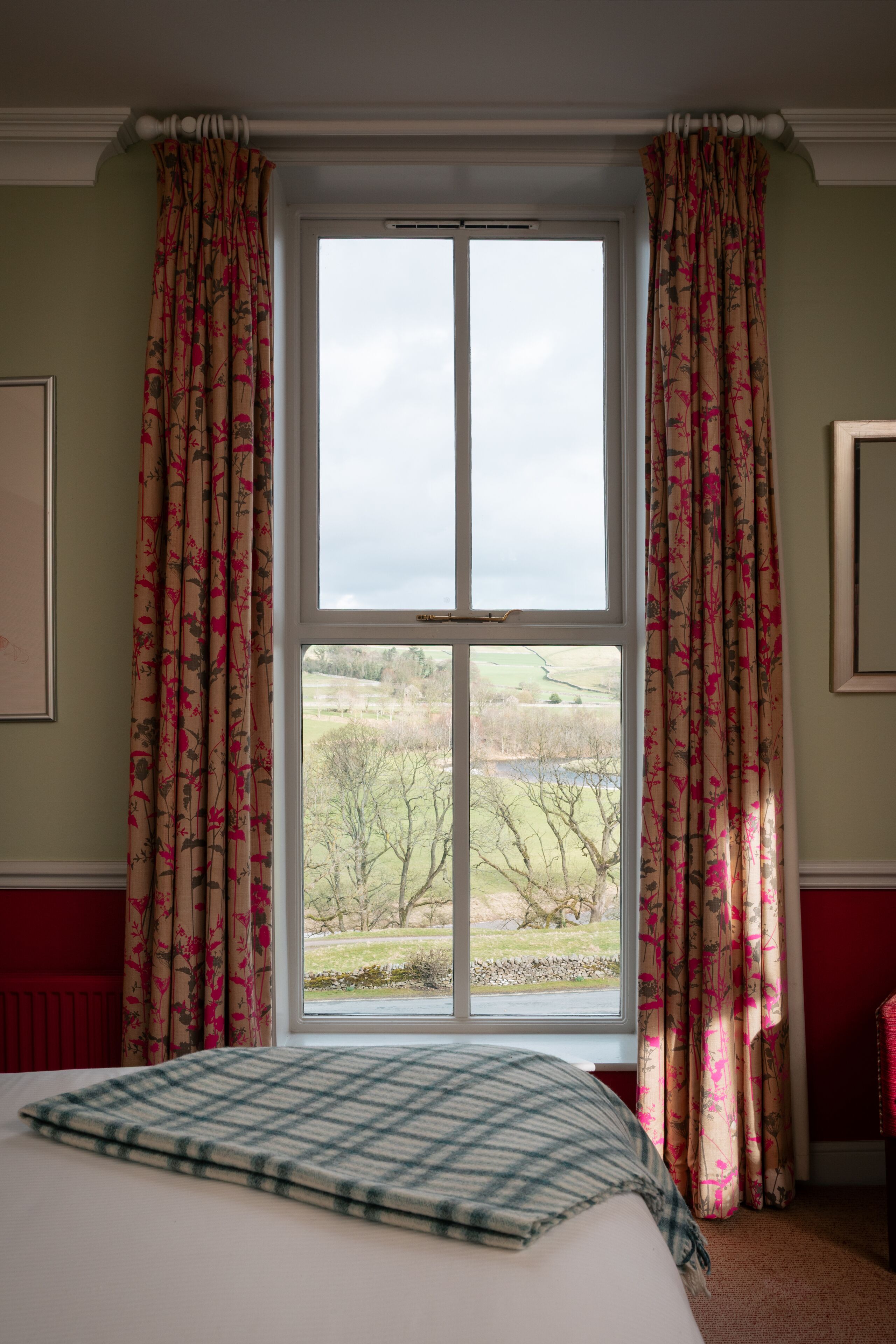 A bedroom with a large window, floral curtains, a plaid bedspread, and a view of a rural landscape.