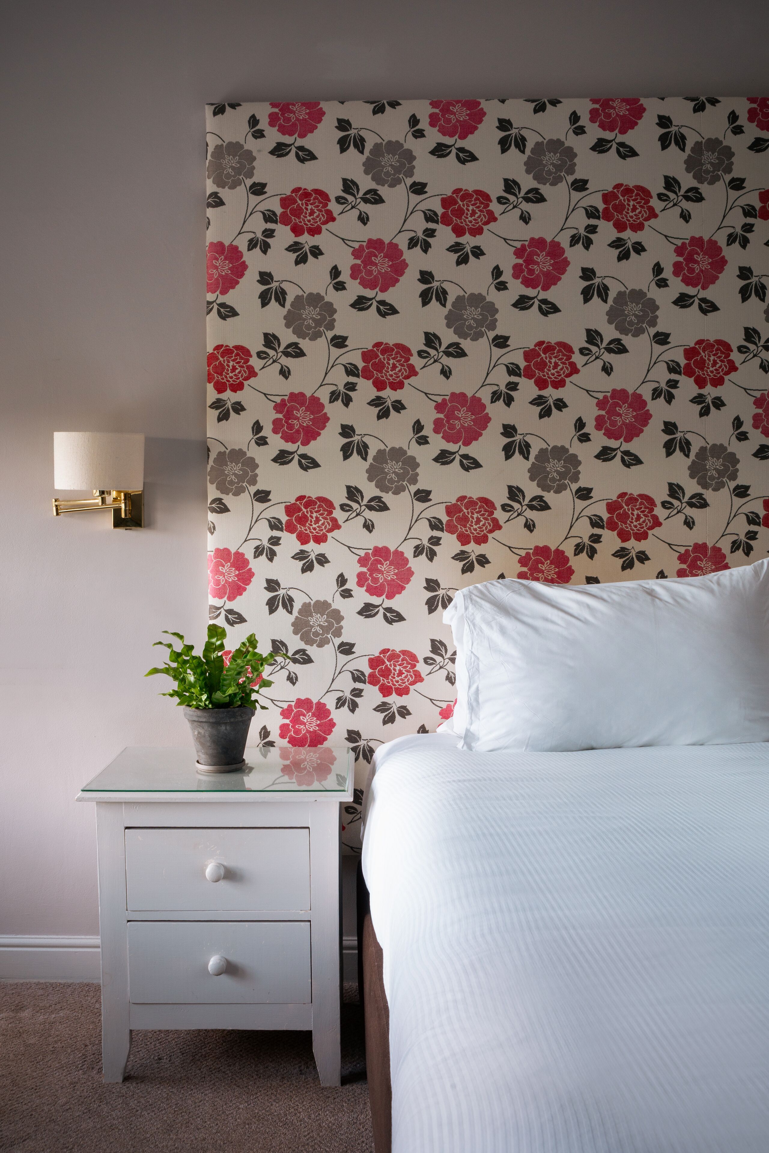 A bedroom with a floral wallpaper, a bedside table with a plant, and a wall-mounted lamp.
