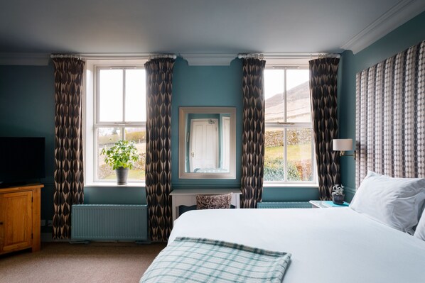A bedroom with a large bed, a window with curtains, a radiator, a television, and a plant. - The Fell Hotel (Skipton)