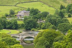 A large house with a stone bridge over a river, surrounded by lush greenery and sheep grazing. - The Fell Hotel (Skipton)