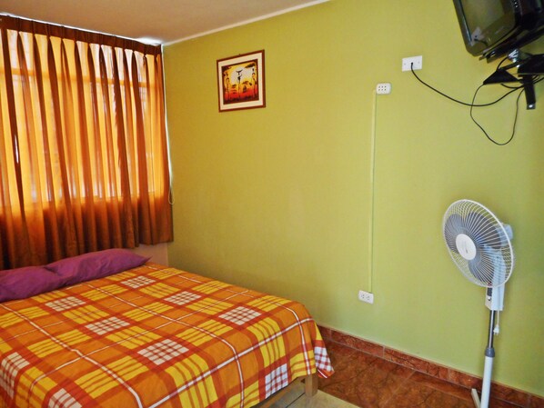 A room with a bed, a standing fan, a television mounted on the wall, and a window with curtains.