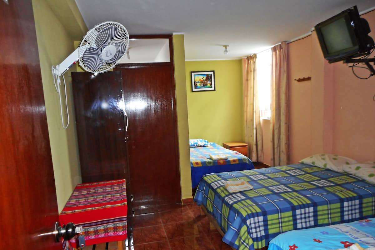 Una habitación con dos camas, una puerta de madera, una mesa con mantel colorido, un ventilador de pie y un televisor en la pared.
