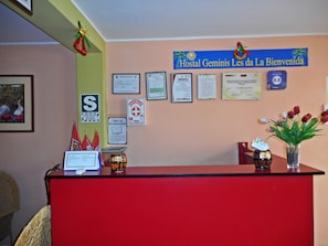 A reception desk with a red counter, a framed certificate, and a sign that reads "Hostal Gemini Les da La Bienvenida".