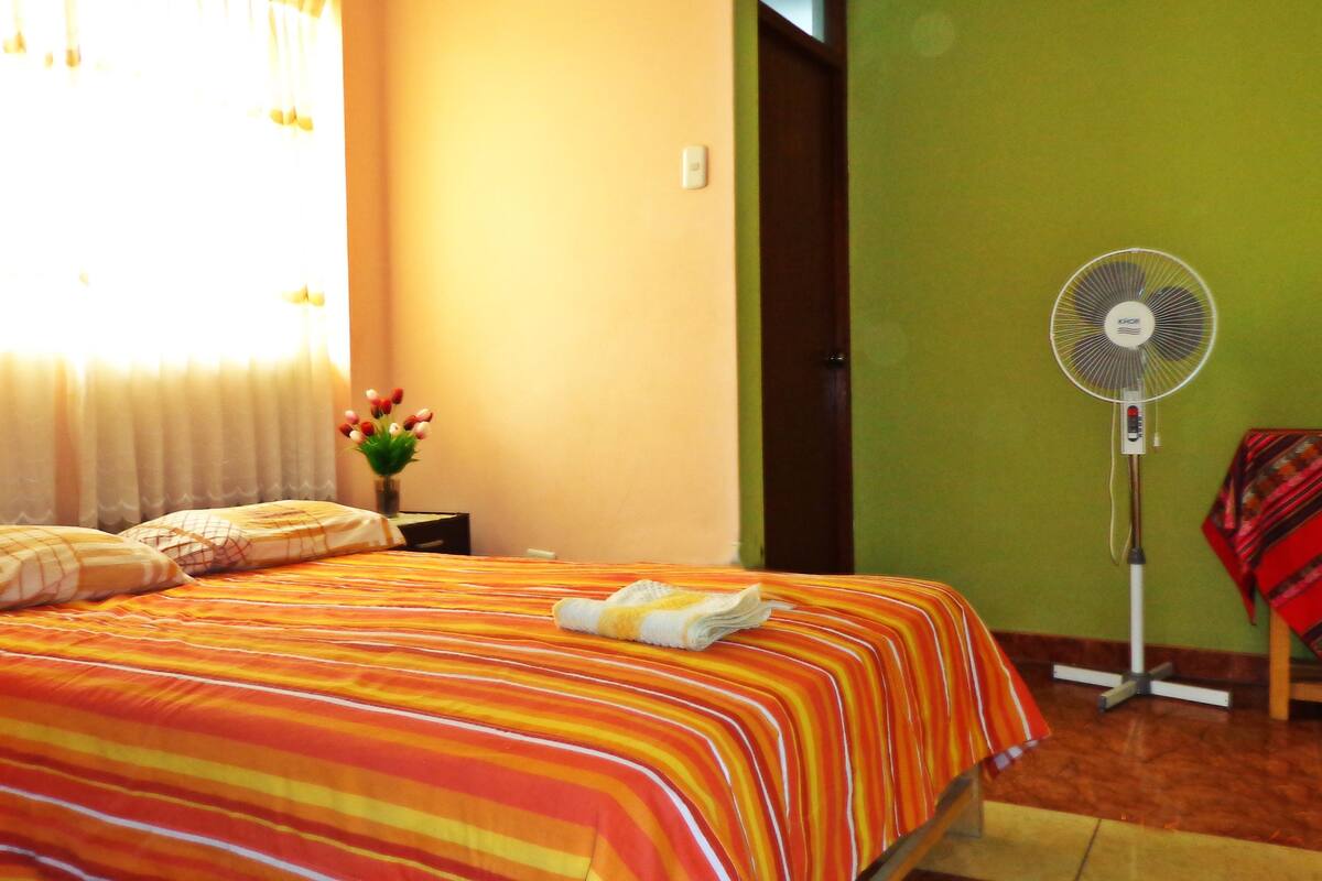 Una habitación de hotel con una cama, una mesita de noche con un florero, un ventilador de pie y una manta roja y blanca a rayas sobre una silla.