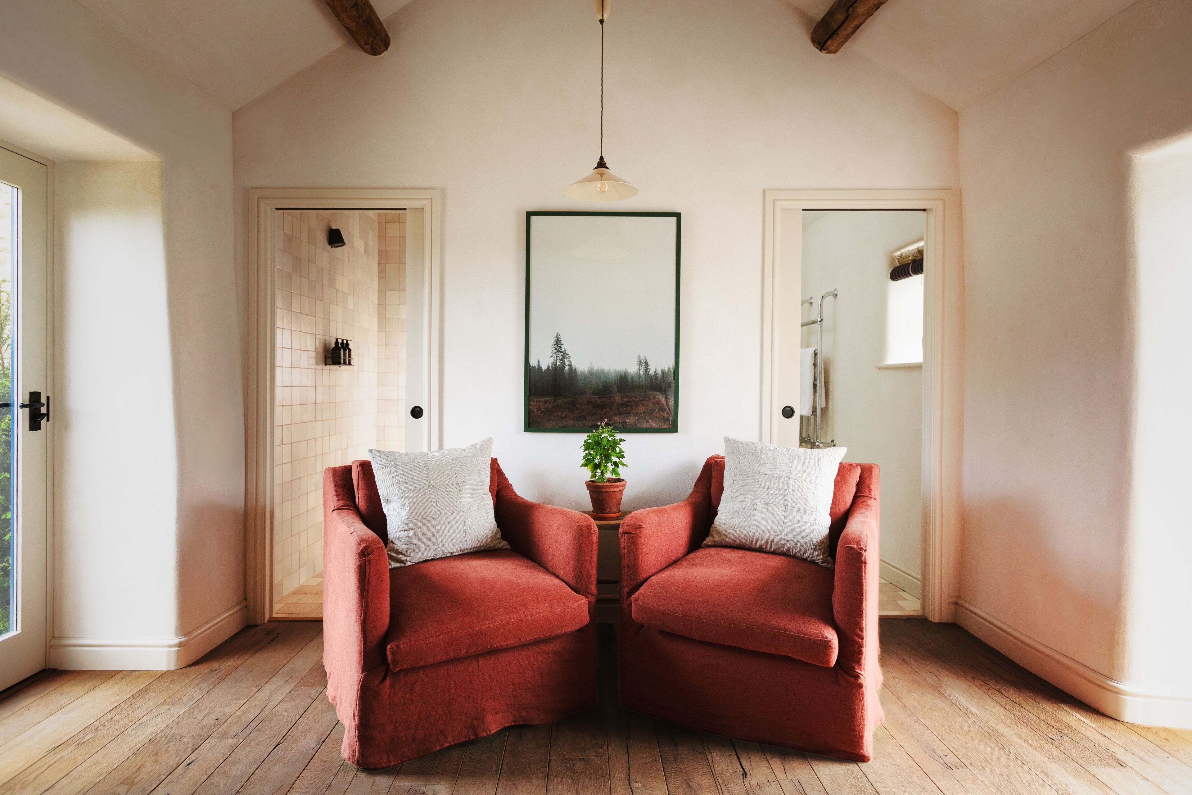 Una habitación con piso de madera, dos sillones rojos con cojines blancos, una planta en maceta, un cuadro en la pared y una puerta con un perchero para toallas.