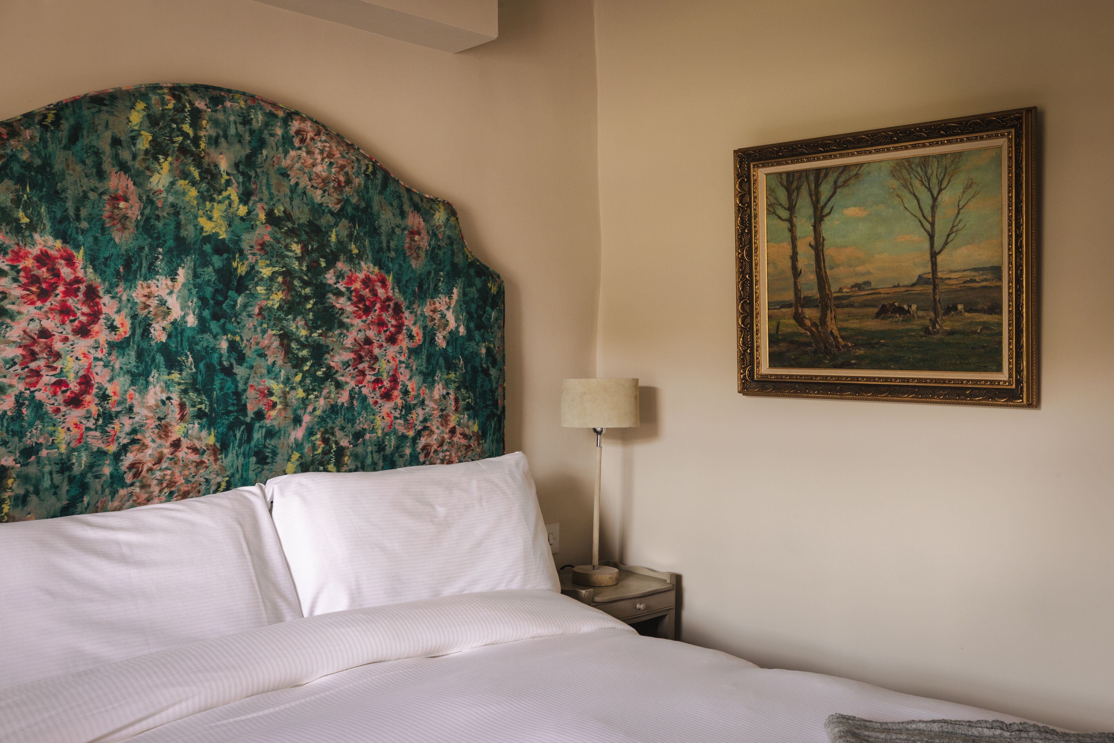 Una cama bien hecha con sábanas blancas, un cabecero floral, un cuadro de paisaje enmarcado y una lámpara de noche en una mesita.