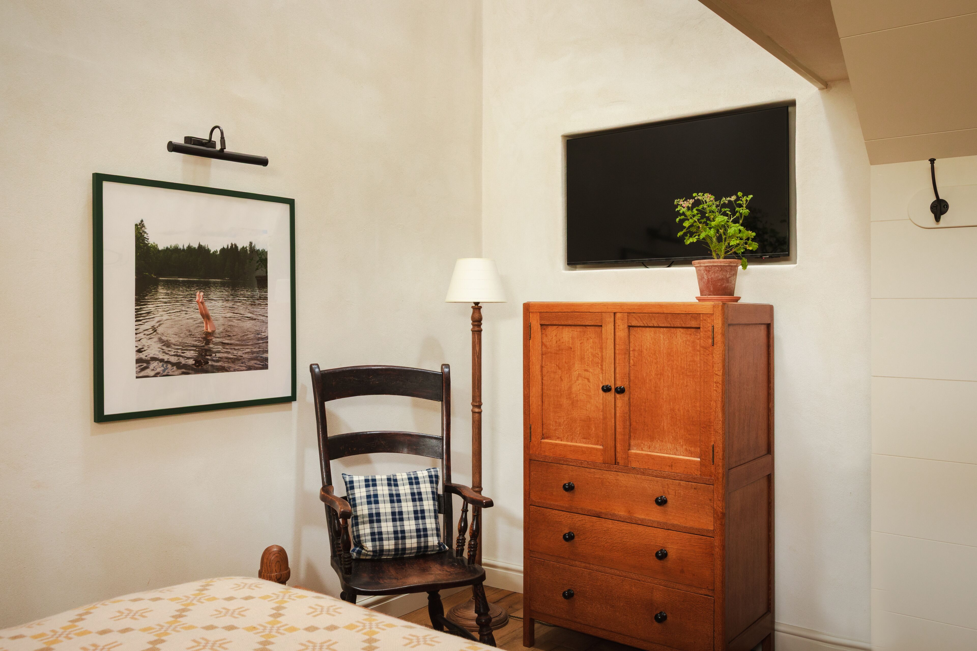 A bedroom with a bed, a wooden chair with a pillow, a wooden cabinet, a framed picture on the wall, and a potted plant on the cabinet.