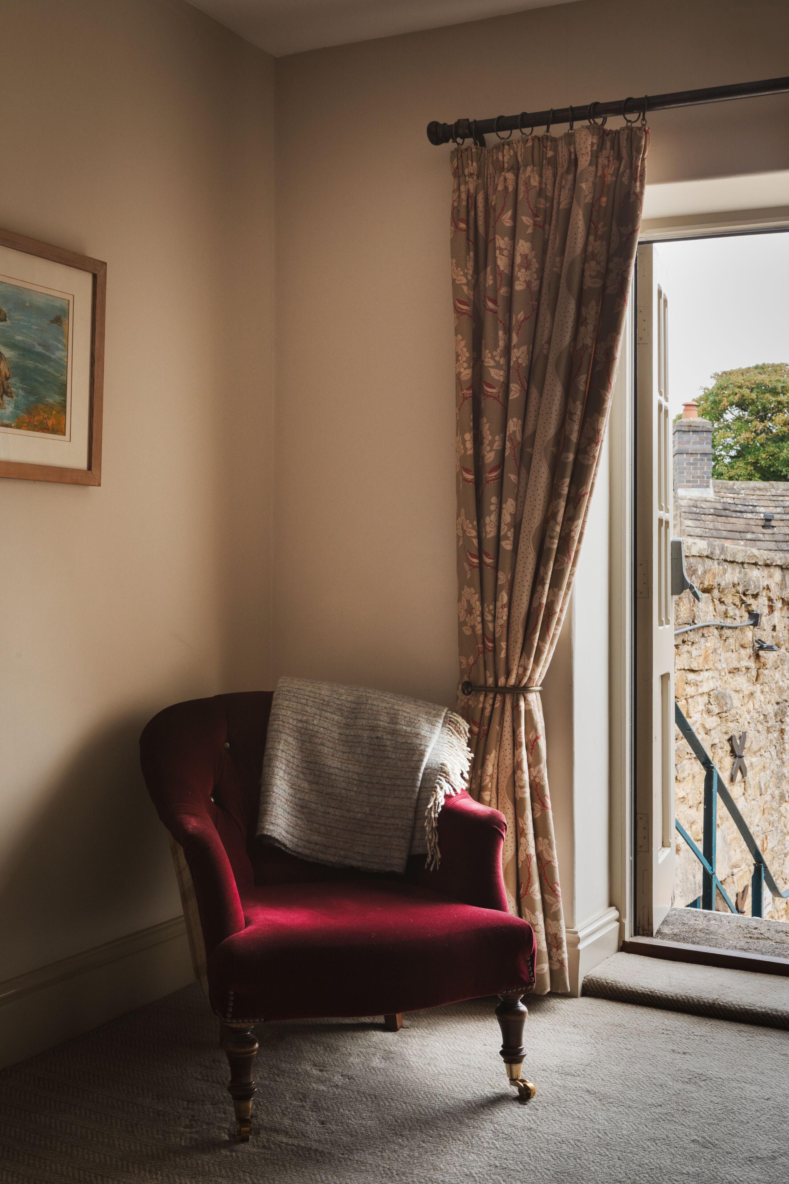 A corner room with a red chair, a window with curtains, a framed picture on the wall, and a stone wall outside.