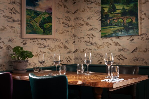 Una mesa de comedor con copas de vino vacías, una planta en maceta y un cuadro de paisaje en la pared.