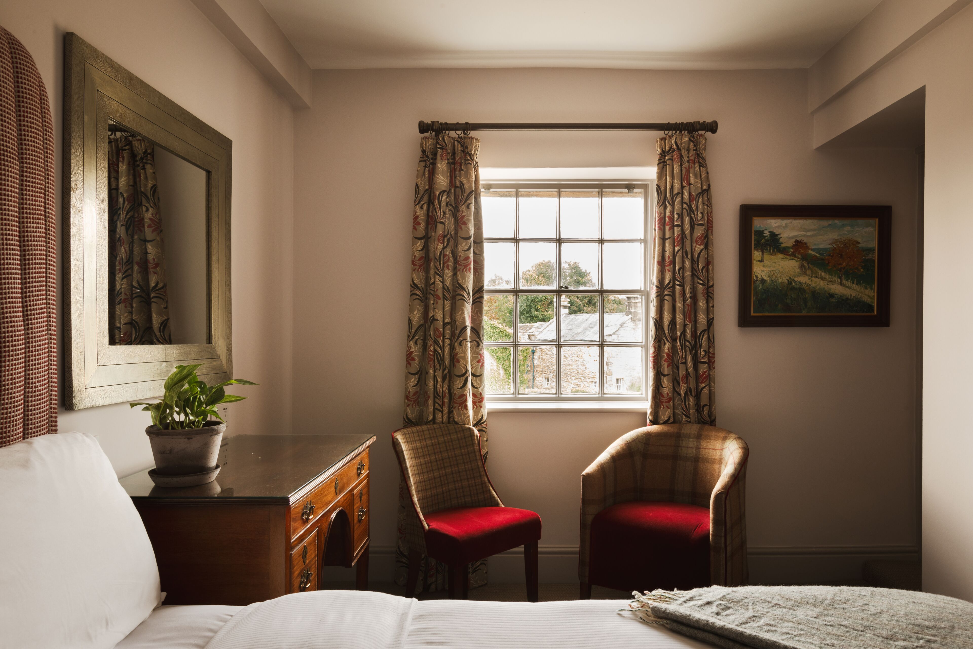 Una habitación con una cama, dos sillas, un aparador, una ventana con cortinas, un cuadro y una planta en maceta.