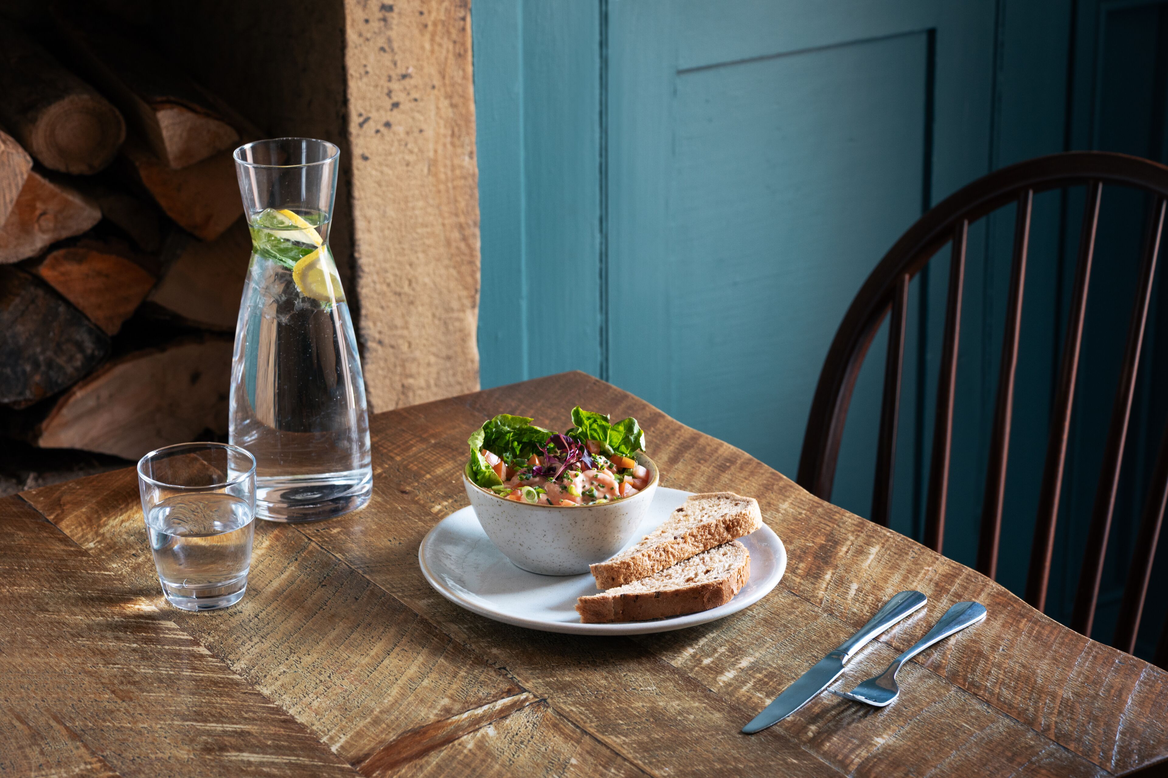 A wooden table with a glass of water, a bowl of salad, and slices of bread on a plate, accompanied by cutlery.