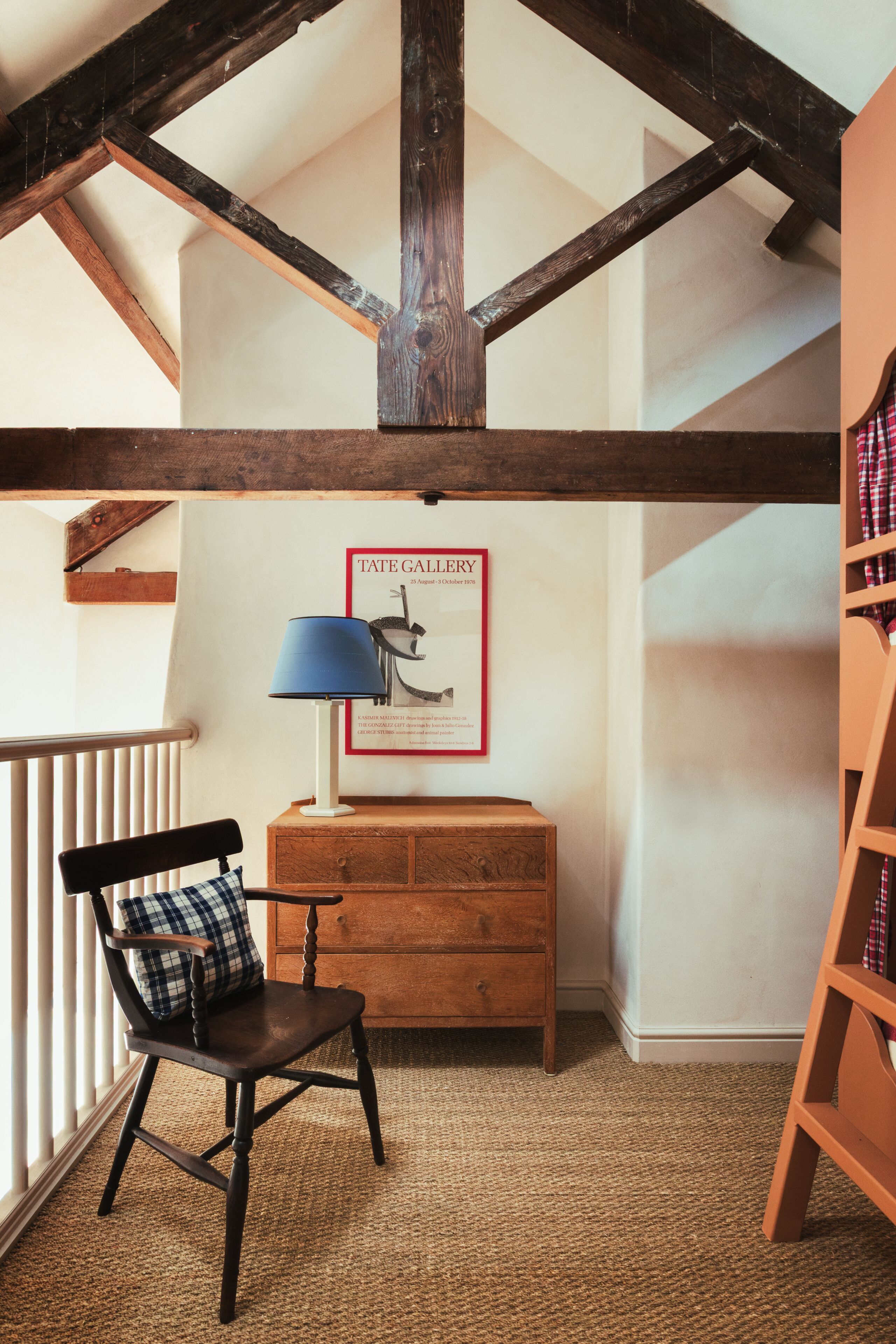 A room with exposed wooden beams, a wooden desk with a lamp, a chair, a framed picture on the wall, and a bookshelf.