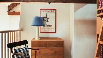 A room with exposed wooden beams, a wooden desk with a lamp, a chair, a framed picture on the wall, and a bookshelf.