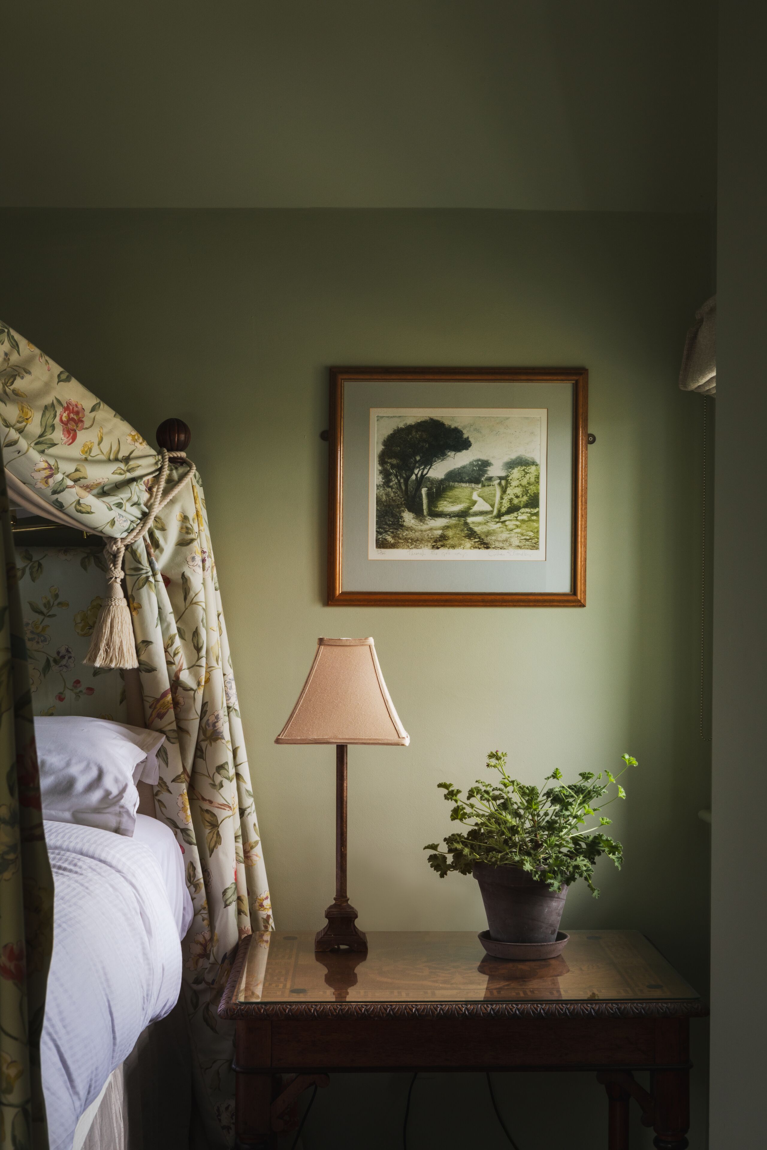 Una habitación con una cama, un cuadro en la pared, una lámpara y una planta en una mesita de noche.