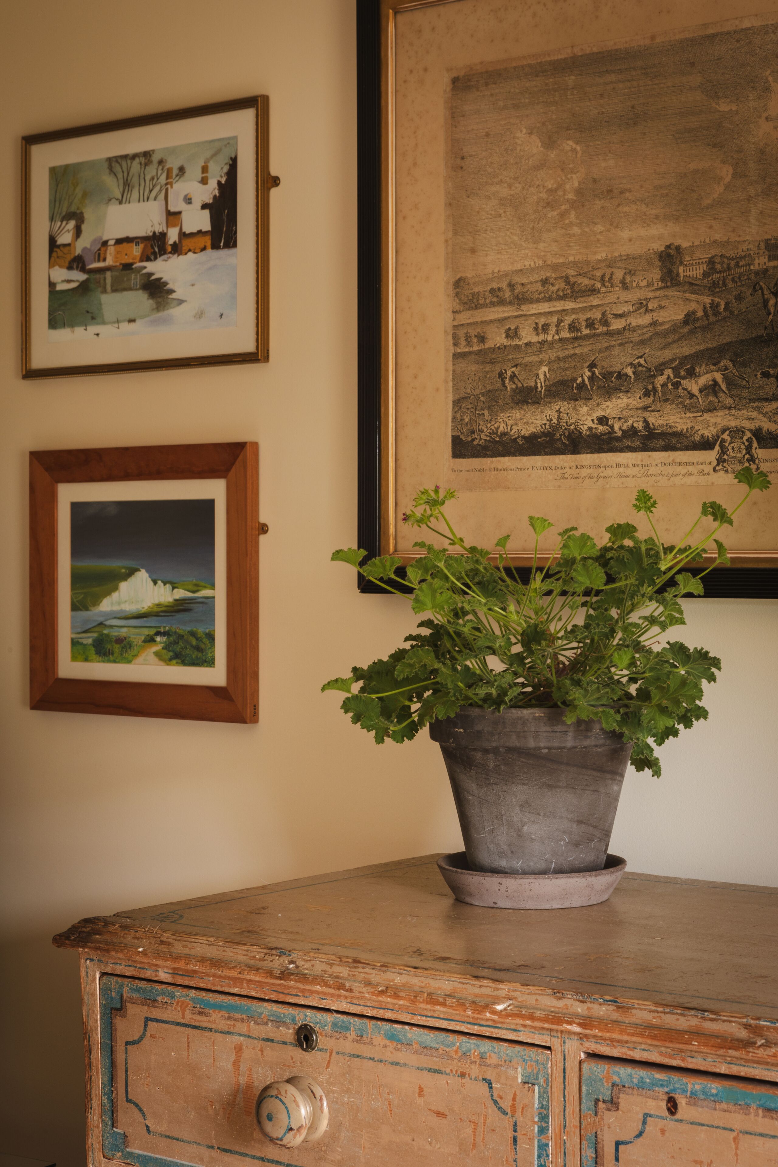 A vintage dresser with a potted plant, three framed artworks on the wall, and a wooden cabinet with a drawer.