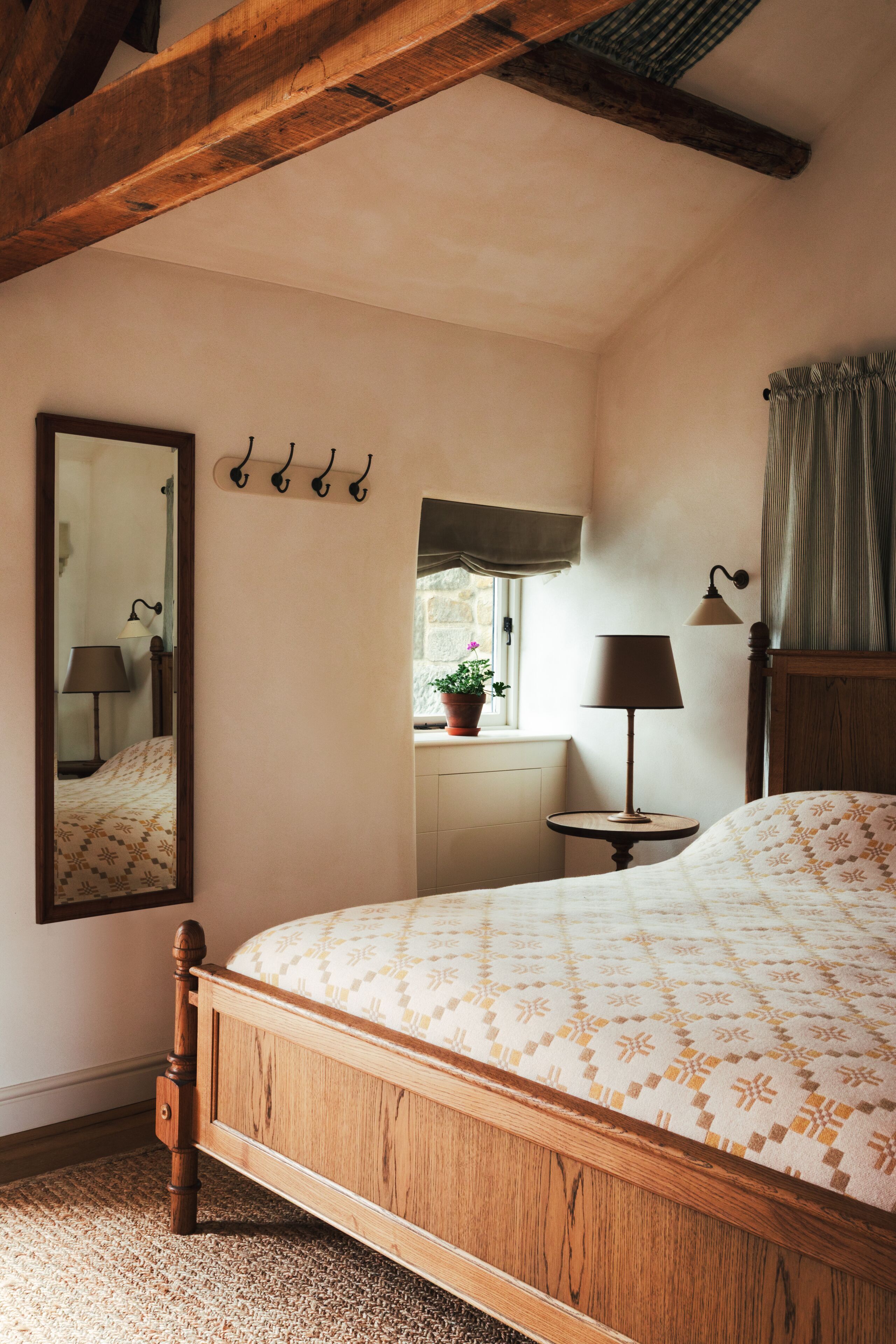 Un dormitorio con cama de madera, espejo, mesita de noche con lámpara, ventana con vista al exterior y un aplique en la pared.
