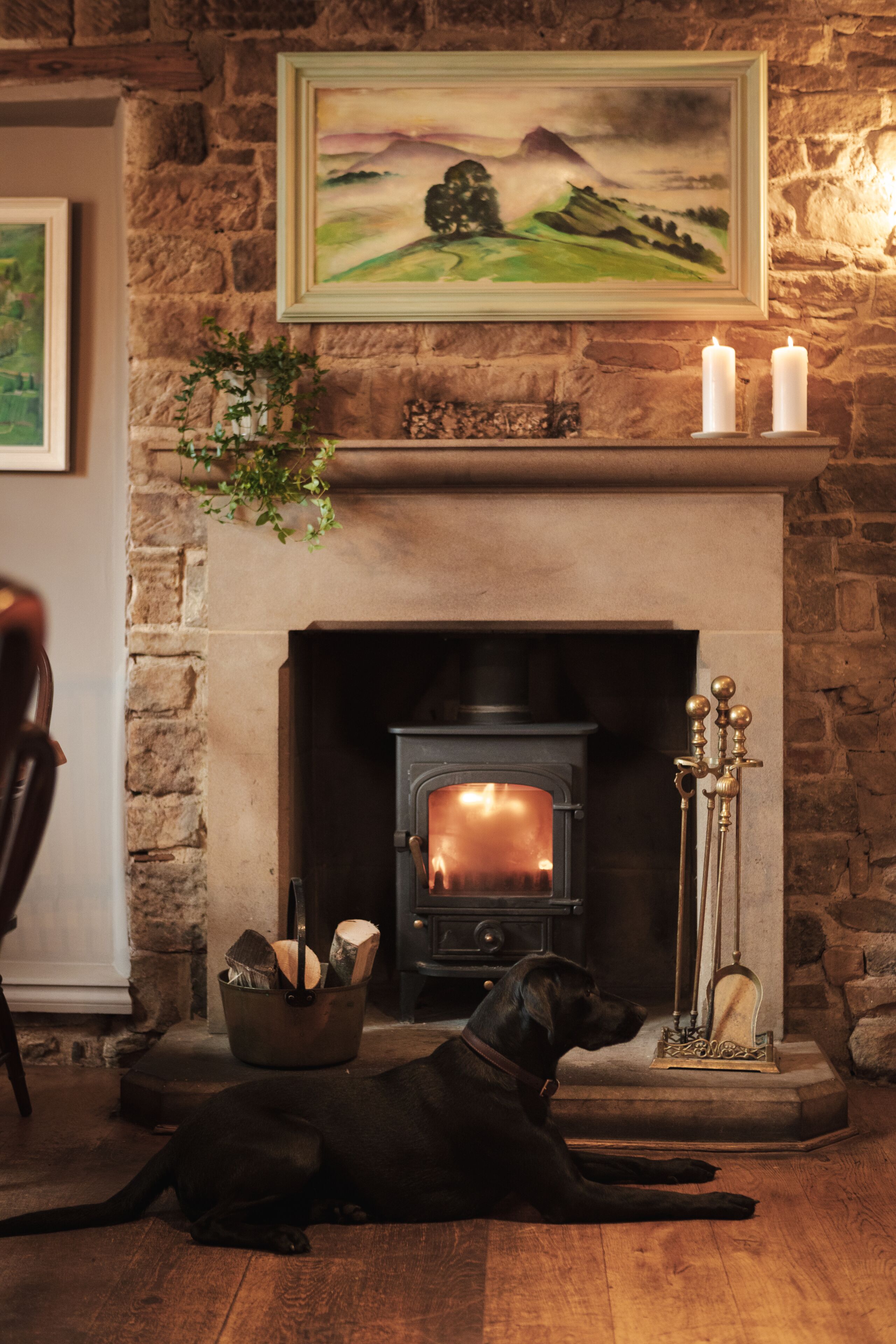 A fireplace with a dog lying in front of it, a metal bucket with logs, and a candle holder on the mantel.