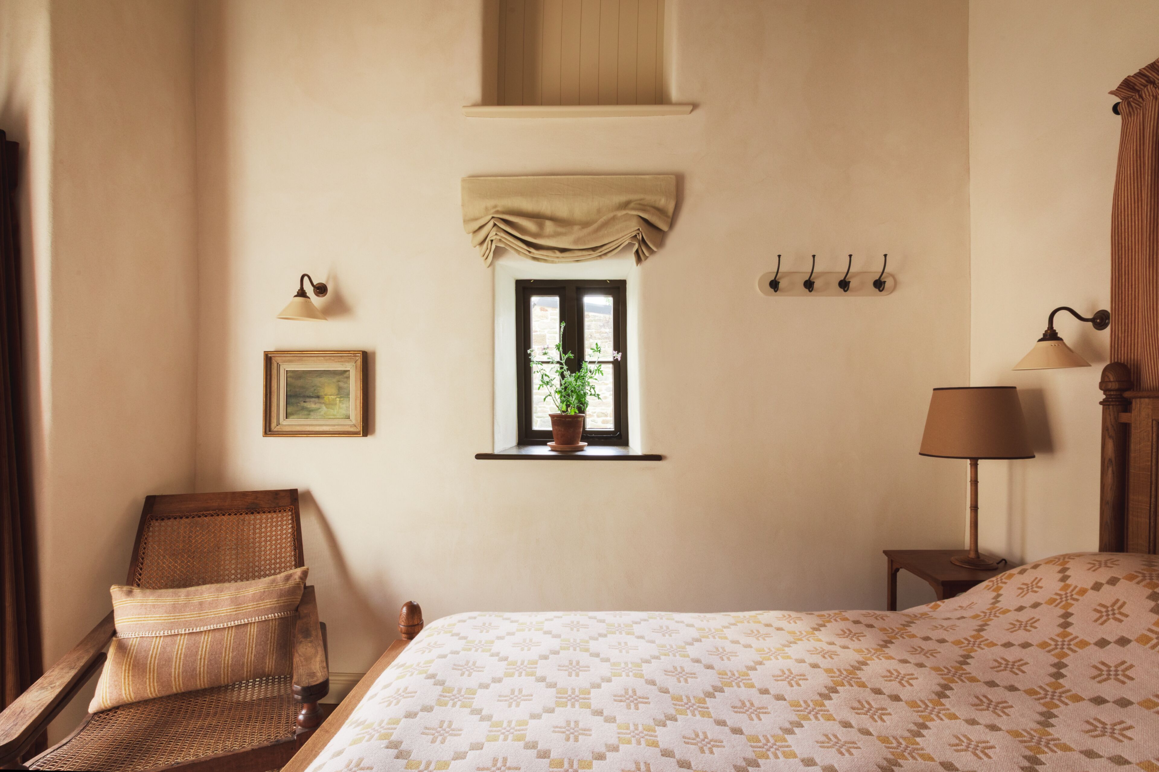 A bedroom with a bed, a chair, a window with a plant, a wall-mounted lamp, and a picture.