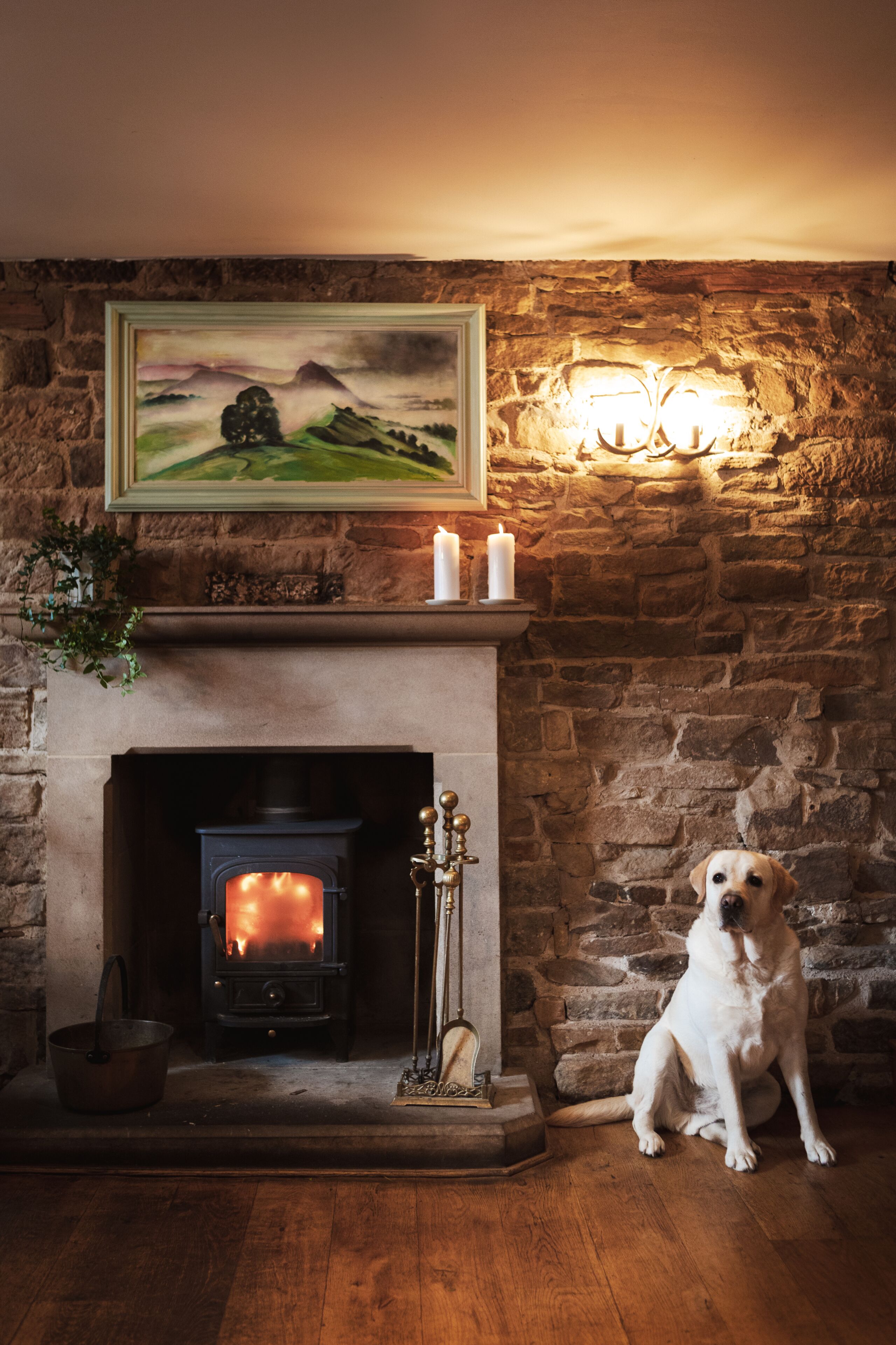 A fireplace with a painting above it, a dog sitting beside the fireplace, and a candle holder on the mantel.
