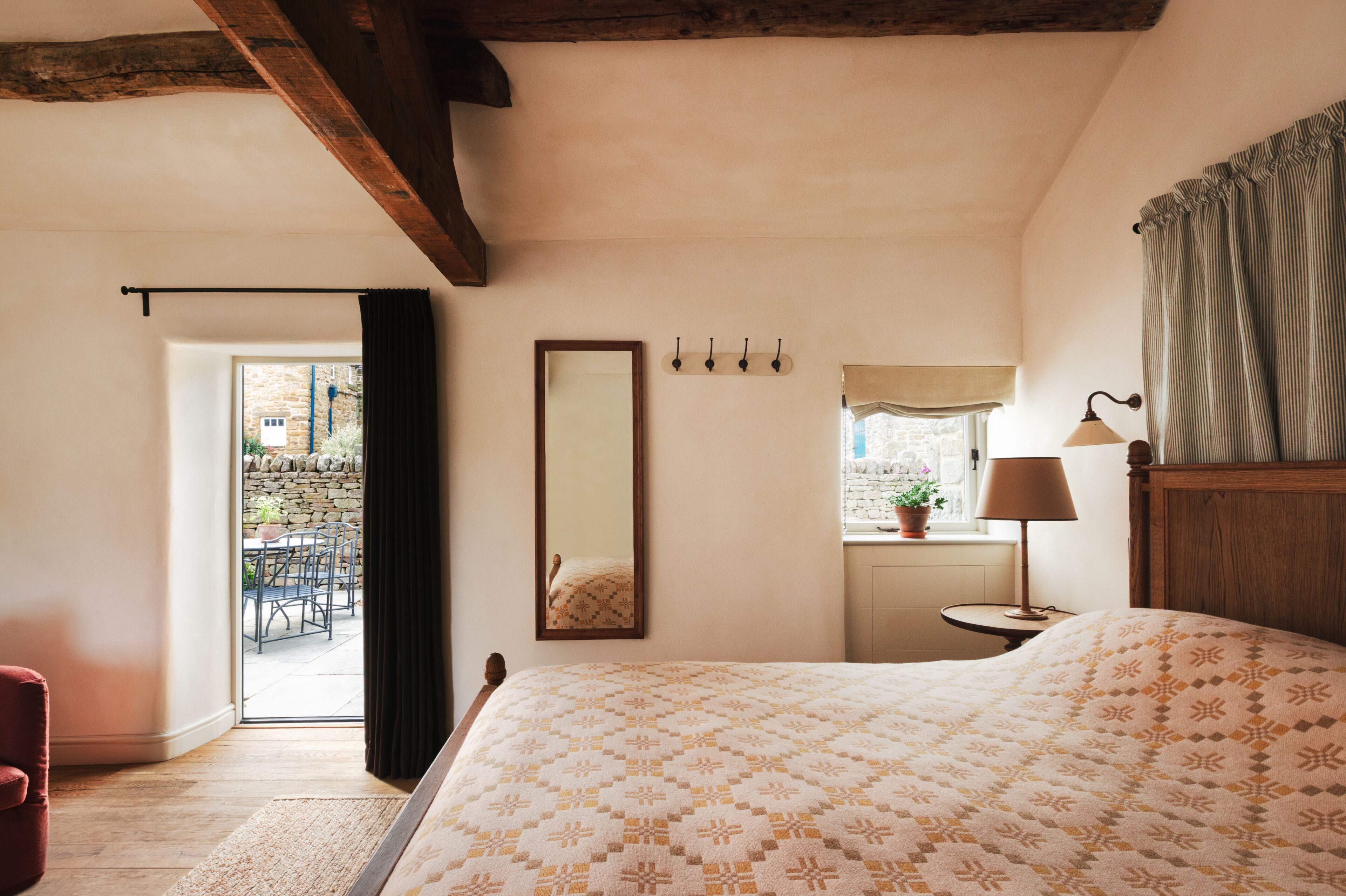 A bedroom with a bed, a bedside table with a lamp, a mirror, and a window with a view of the outside.