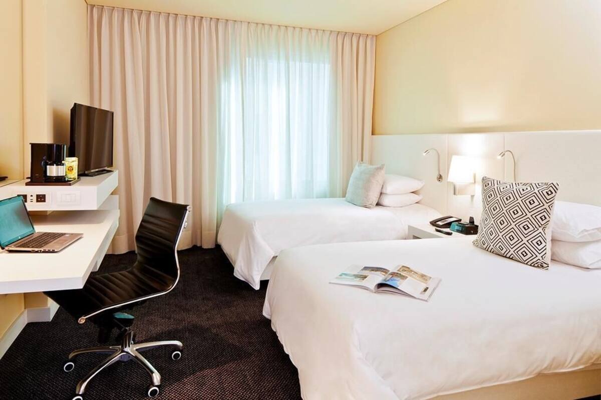 A hotel room with two beds, a desk with a laptop, a chair, a TV, and a window with curtains.