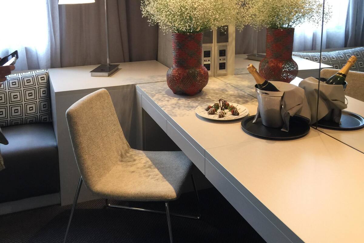 A modern hotel room with a large mirror, a desk with a chair, a vase with flowers, and a bottle of champagne.