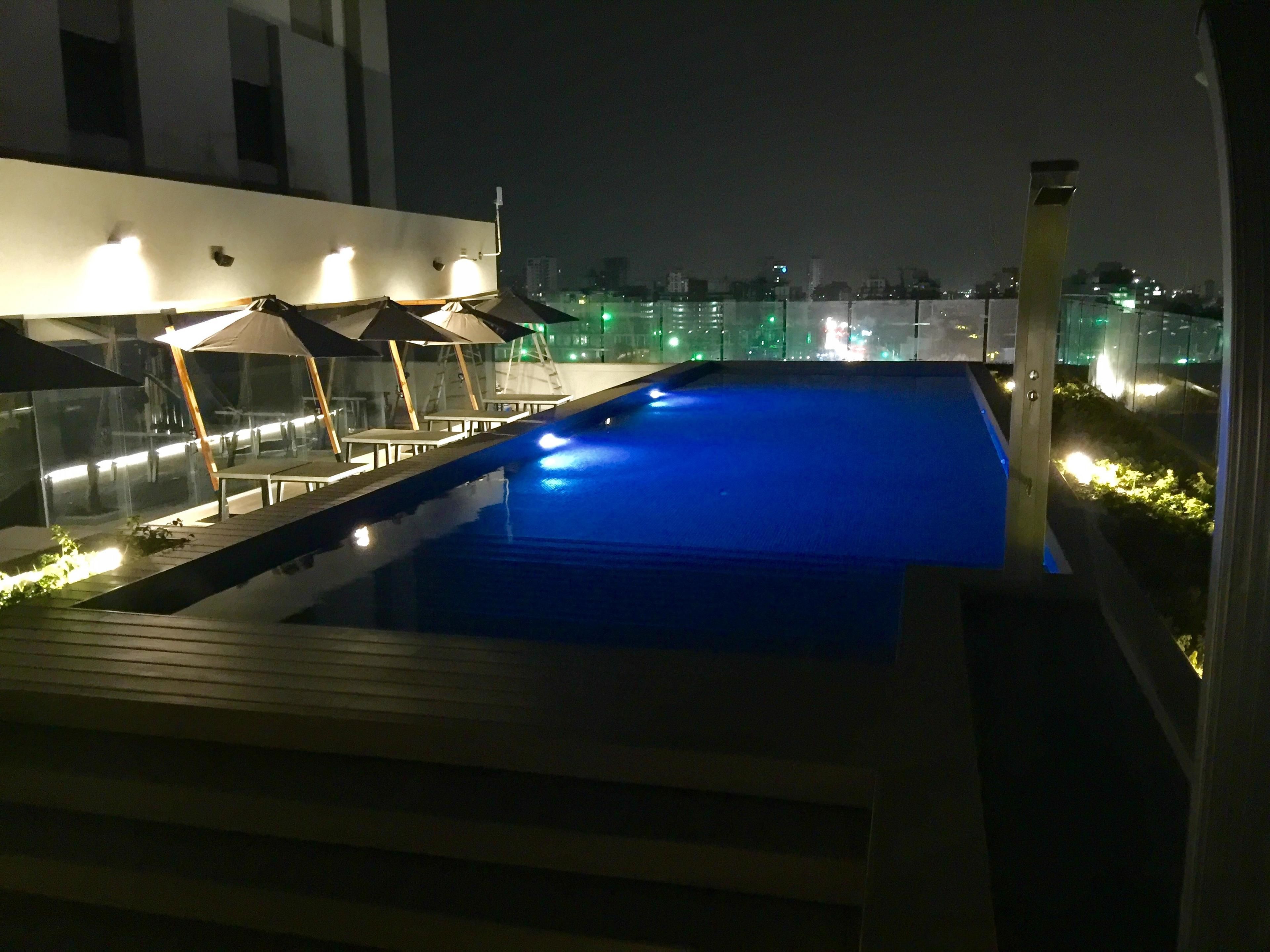 A rooftop pool with a blue water feature, surrounded by lounge chairs and umbrellas, with city lights in the background.