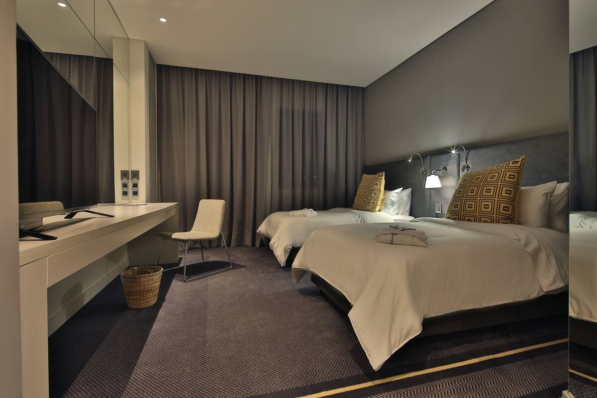 A modern hotel room with a desk, two beds, a chair, and a large mirror.