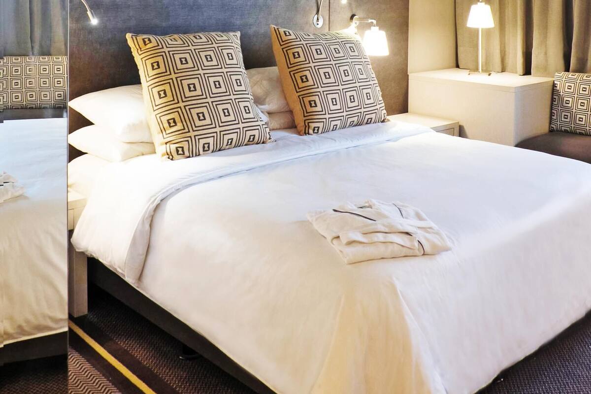 A hotel room with a large bed, a headboard with integrated lighting, a bedside table with a lamp, and a view of the sea through the window.
