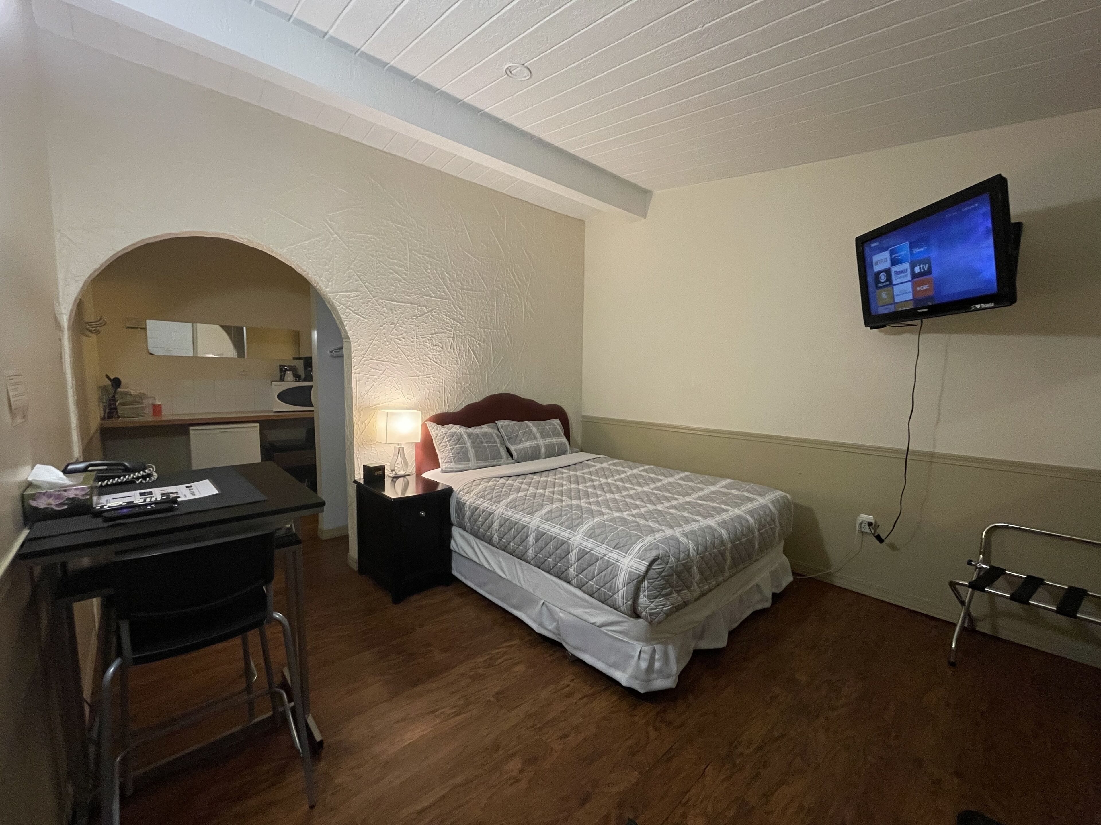 A bedroom with a bed, a desk, a chair, a TV mounted on the wall, and a small kitchenette area.
