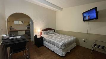 A bedroom with a bed, a desk, a chair, a TV mounted on the wall, and a small kitchenette area.
