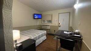 Habitació petita i ben il·luminada amb un llit, escriptori amb cadira, televisió penjada a la paret i una porta.