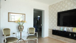 Ein Wohnzimmer mit zwei Sesseln, einem kleinen Glastisch mit Blumenvase, einem großen Flachbildfernseher und einer Wand mit Muster