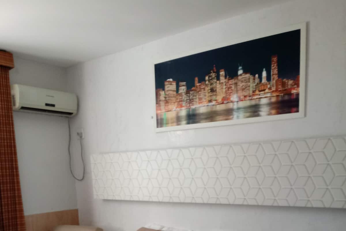 A hotel room with a bed, a chair, a nightstand, a lamp, and a cityscape painting on the wall.