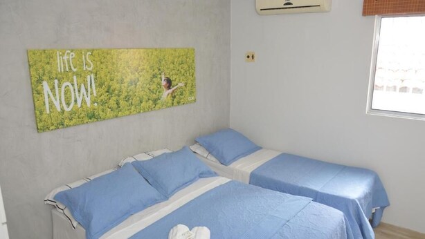 Ein Zimmer mit zwei Betten, jeweils mit blauer Bettwäsche und Kissen, einer Wand-Klimaanlage und einem motivierenden Zitat an der Wand.