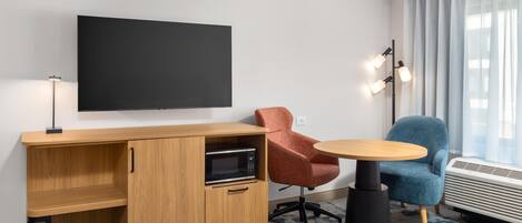 A hotel room with a flat-screen TV, a wooden desk with a microwave, a red chair, a blue chair, and a small round table.