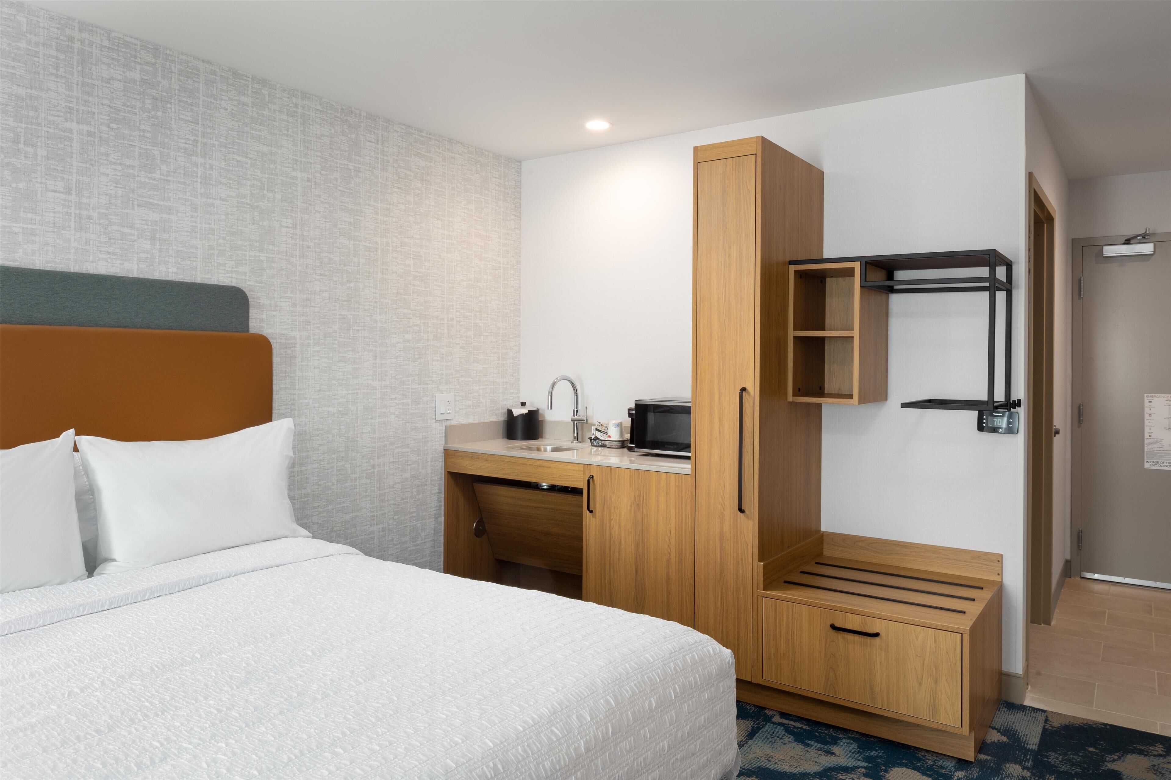 A hotel room with a bed, a wooden desk with a microwave, a sink, and a wardrobe.