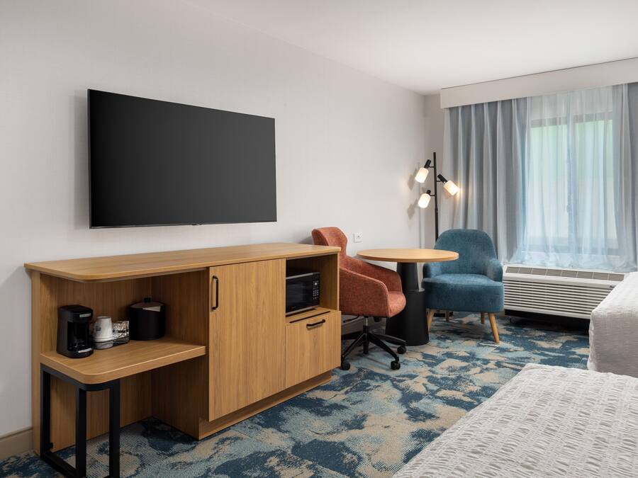 Habitació d'hotel amb un llit, un escriptori amb una cadira, una televisió de pantalla plana, una taula petita, un butacó blau, una làmpada de peu i una finestra amb cortines.