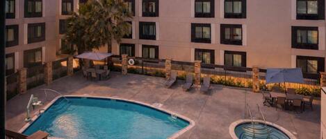 A hotel pool area with a rectangular pool, a smaller circular pool, lounge chairs, umbrellas, and a palm tree.