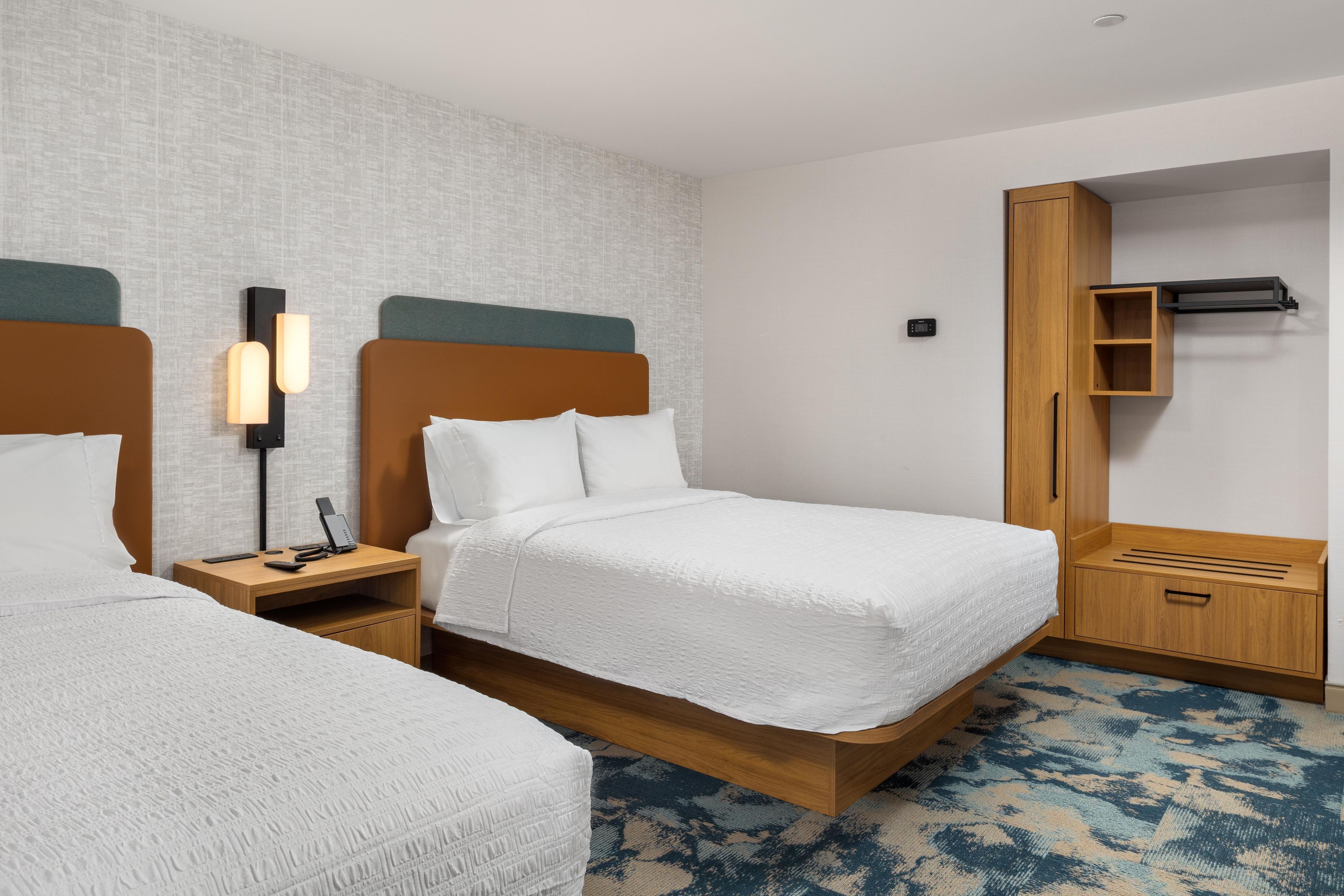A hotel room with two beds, a bedside table with a phone and remote control, a wall-mounted light, and a wardrobe with a mirror.