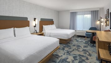 Quarto de hotel com duas camas, uma mesa de trabalho, uma cadeira, luminária de mesa e uma janela com cortinas.
