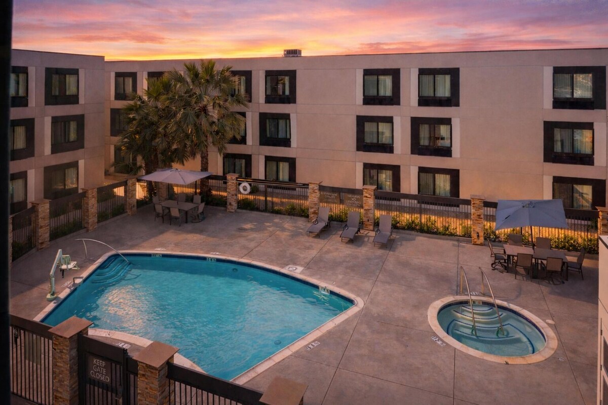 Un área de piscina de hotel con una piscina rectangular, una piscina circular más pequeña, sillas de descanso, sombrillas y una palmera.